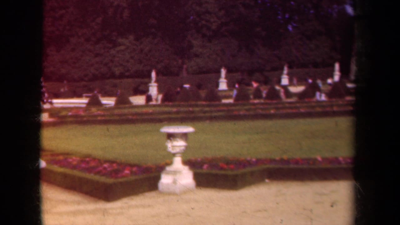 Formal garden with lawn, flowerbeds, stone urn, hedges, trees, and distant visitors.