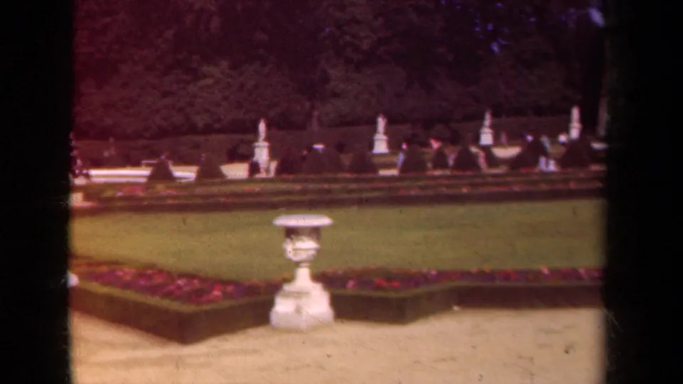 1939 Paris home-movie view of a formal garden with a central stone urn, clipped lawn, flowerbeds, and distant visitors near hedges.