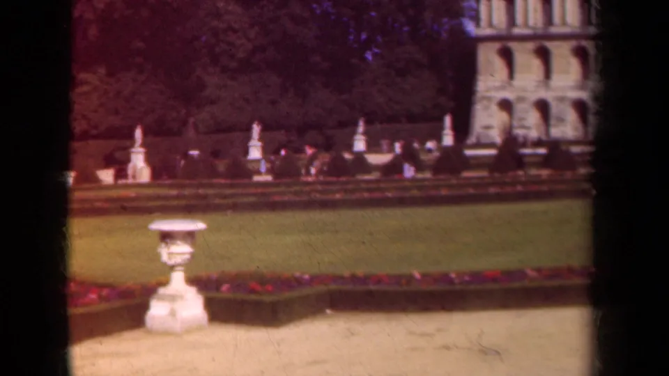 The camera shifts across the Paris garden, showing a stone urn, clipped planting beds, distant visitors, and the edge of a historic facade.