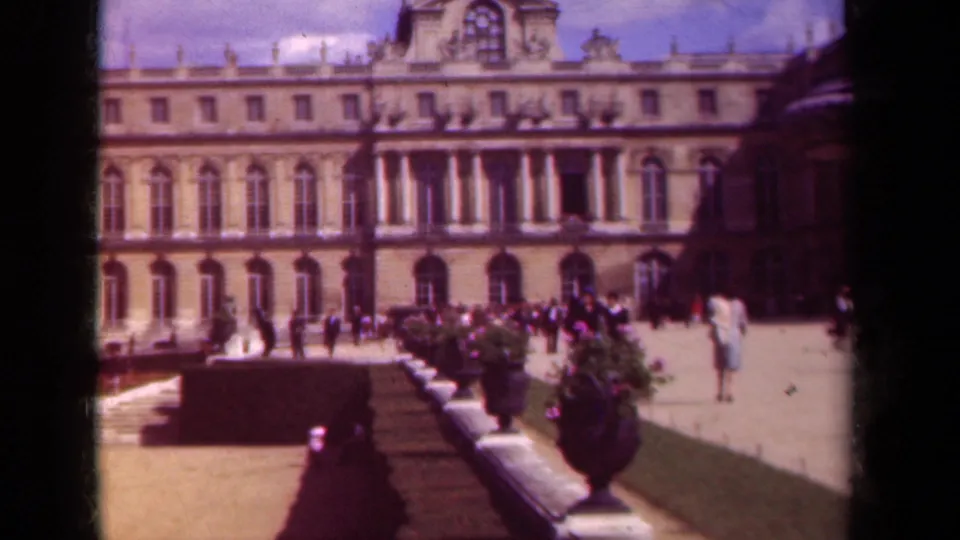 1939 Paris frame of a palace-like exterior seen from a garden path, with visitors in light clothing moving near large planters.