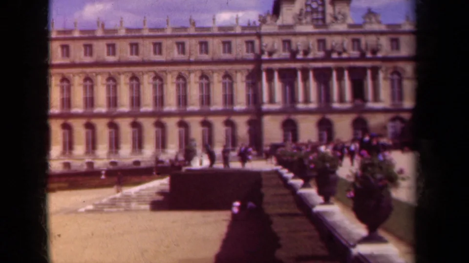 1939 Paris home-movie frame with warm color and soft focus on a historic facade, formal plantings, and distant courtyard visitors.