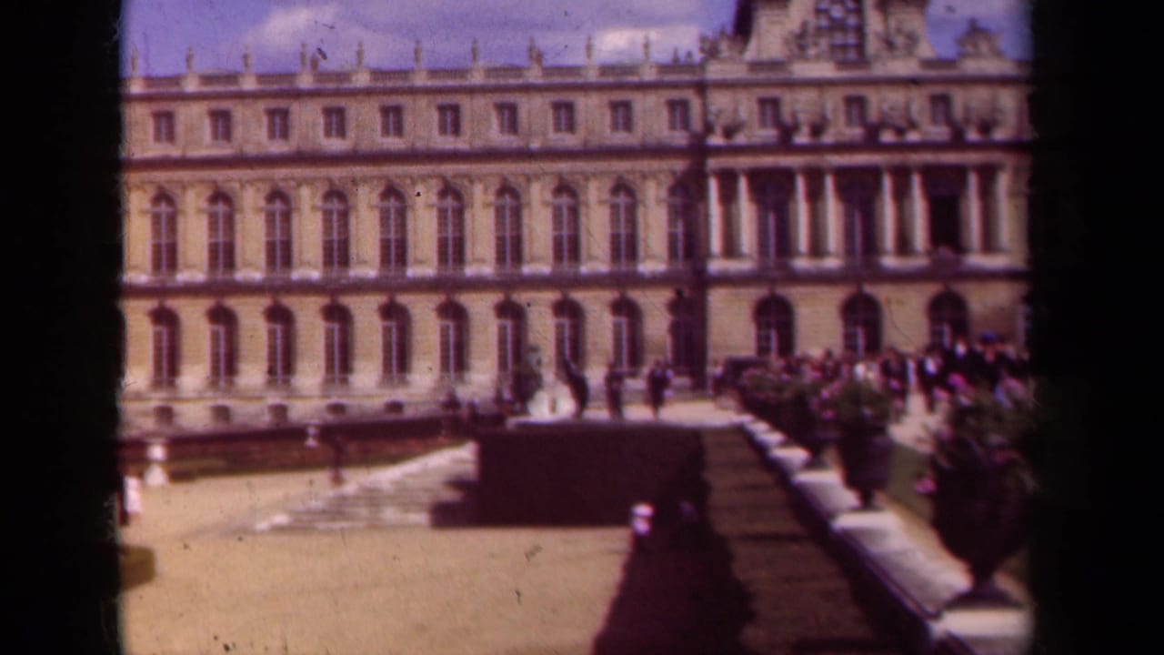 1939 8mm frame of a grand Paris-area palace courtyard, with an ornate facade, hedges, steps, potted plants, and distant visitors.
