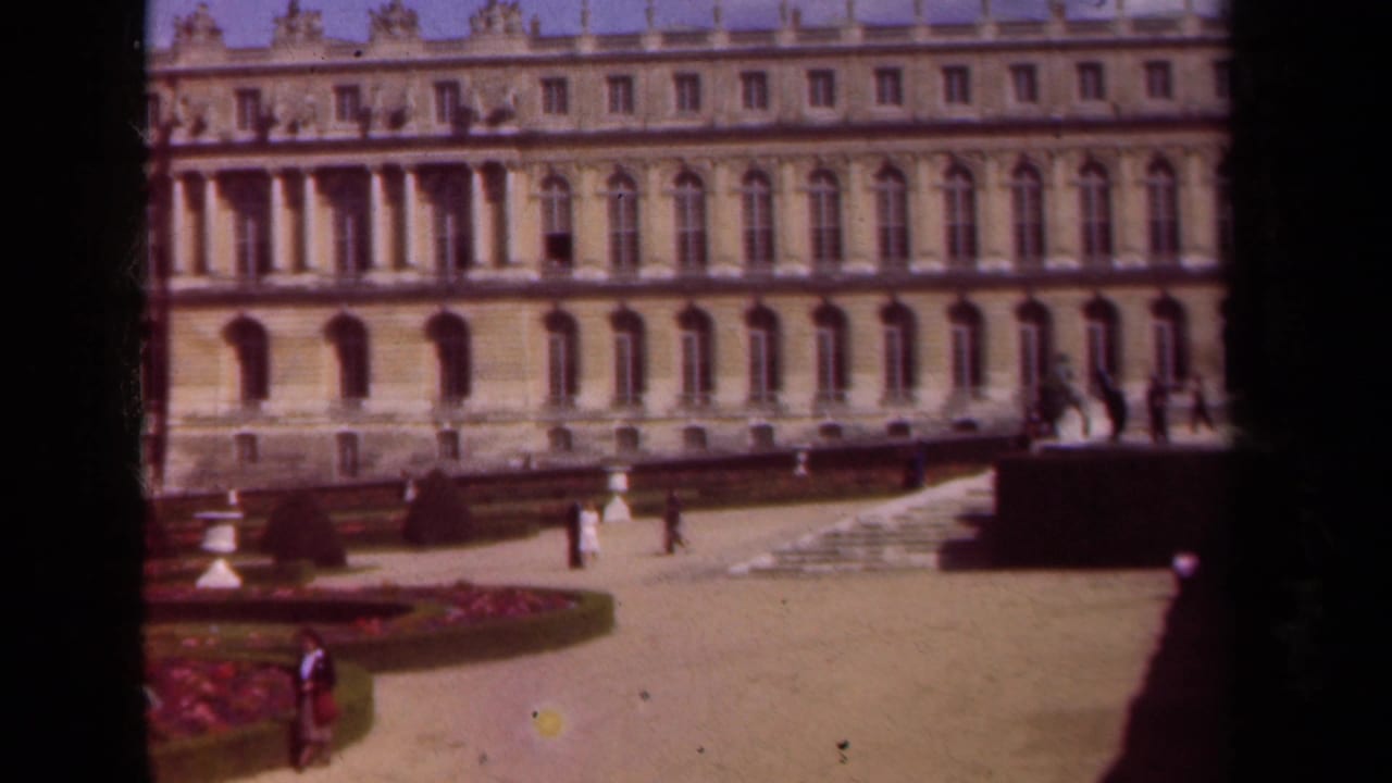 Ornate palace facade beyond a courtyard and garden beds with a few visitors walking.