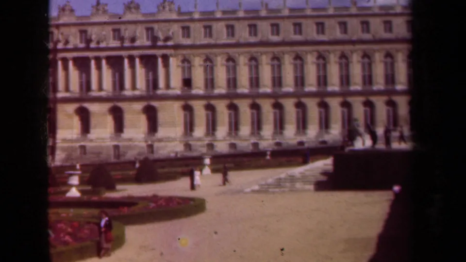 1939 Paris home-movie view of a grand palace facade, courtyard paving, garden beds, and scattered visitors in sunlight.