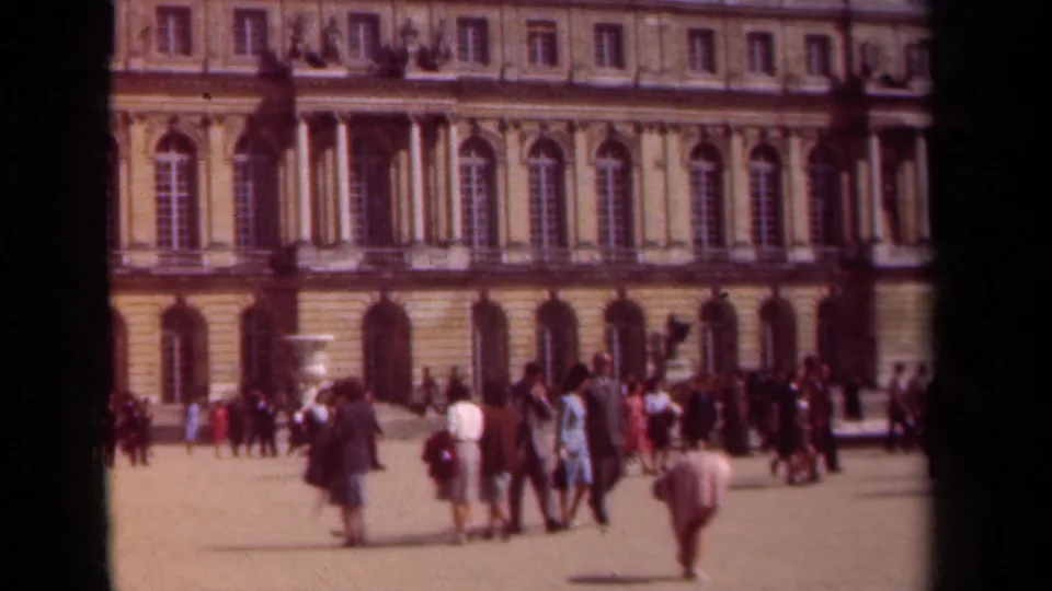 1939 Paris footage frame with tourists in coats and hats before a formal palace facade and open courtyard.