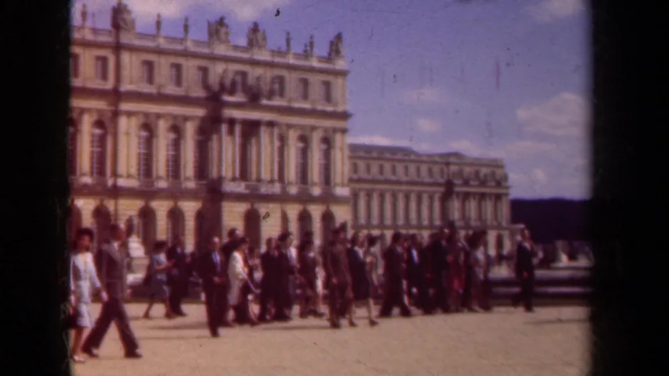 A large group of visitors in period clothing walks across a Paris palace courtyard, with columns and statues visible behind them.