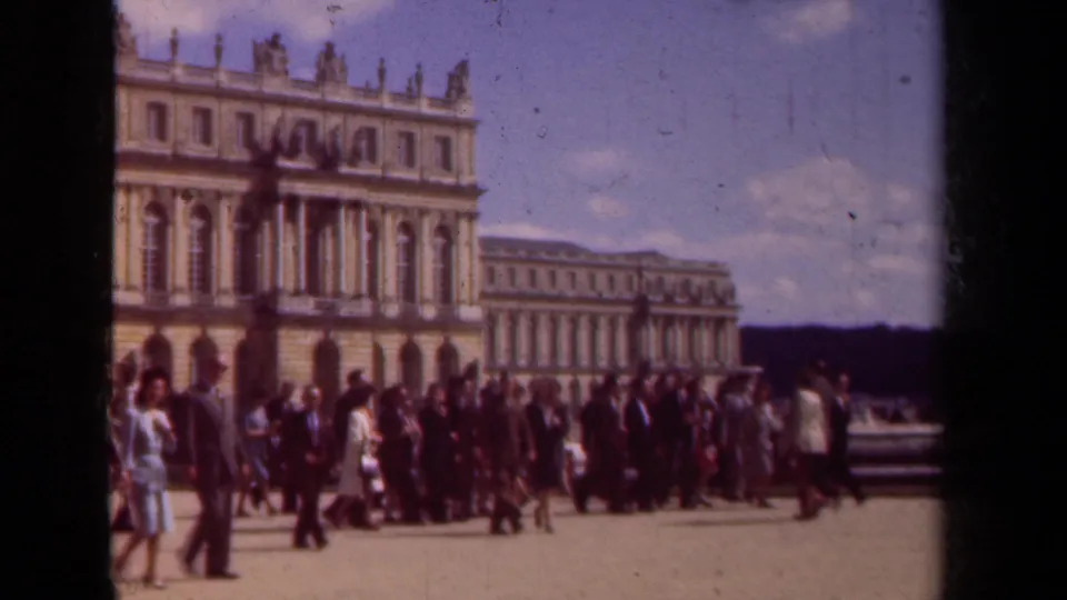 The frame shows tourists clustered in a formal courtyard in Paris, with a historic palace facade and bright clouds in view.