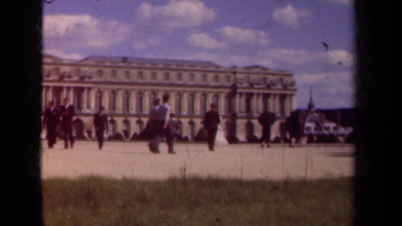 Tourists walk across a palace courtyard with a long stone facade behind them.