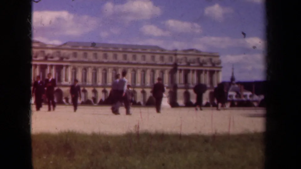1939 8mm view of tourists crossing a broad courtyard before a grand palace facade near Paris, France.