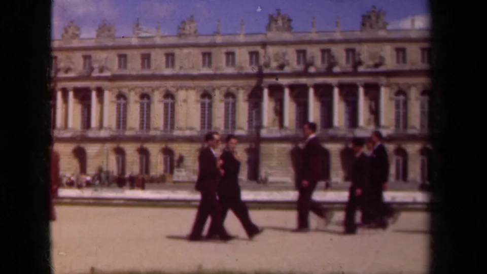Adults in dark suits gather and pass through a Paris palace courtyard, with ornate architecture behind them on 8mm film.