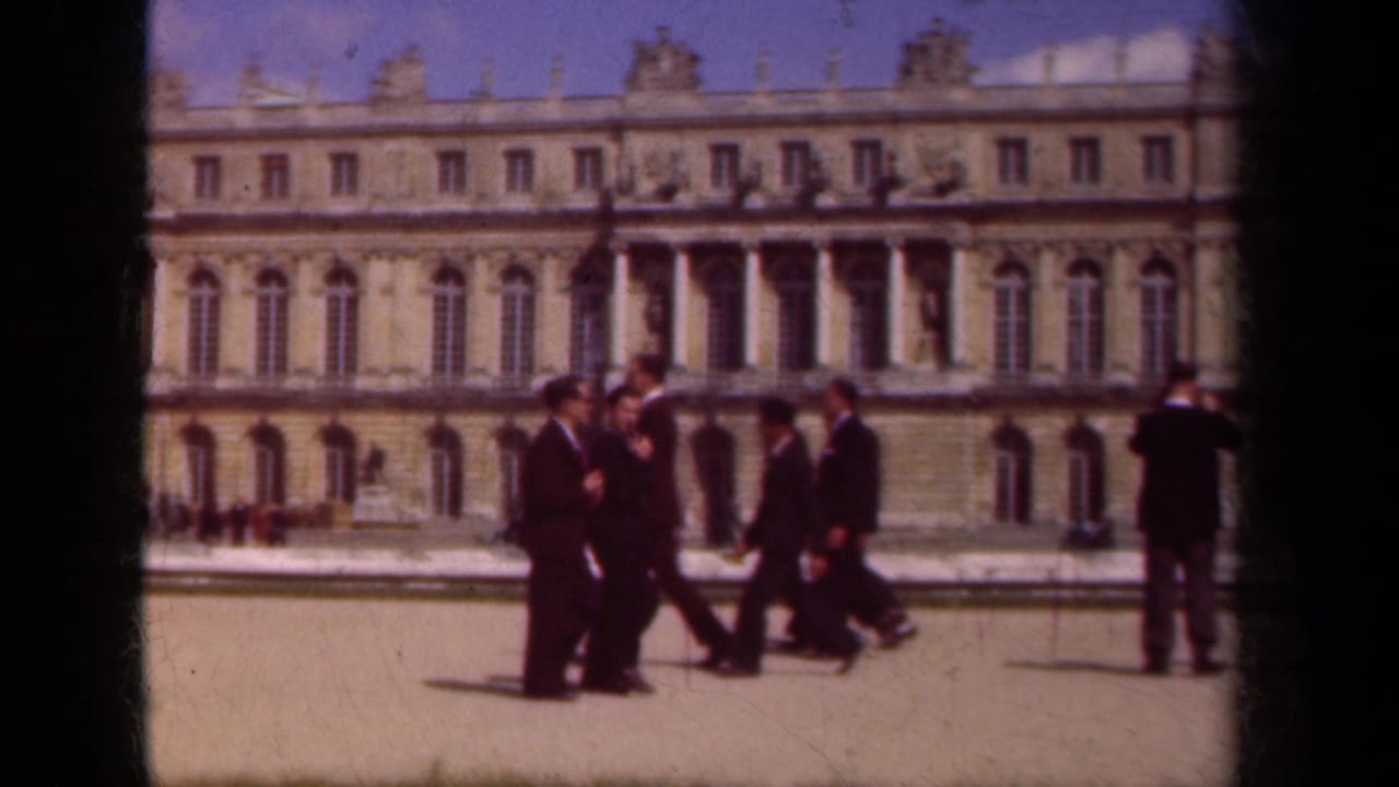 In Paris, France, 1939, suited men walk through a bright palace courtyard with columns, arched windows, and roofline sculpture behind them.