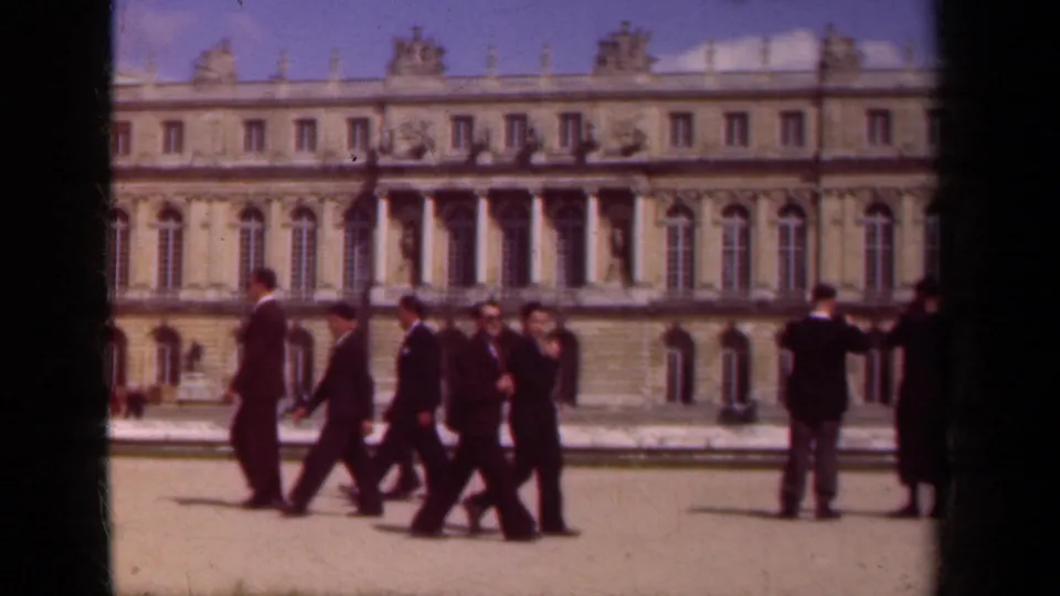 Suited men move across a Paris palace courtyard in 1939, framed by classical stone architecture and distant visitors.