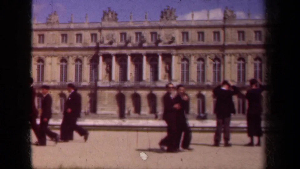 Visitors stand and walk in a Paris palace courtyard in 1939, with a large stone facade and classical ornament in the background.