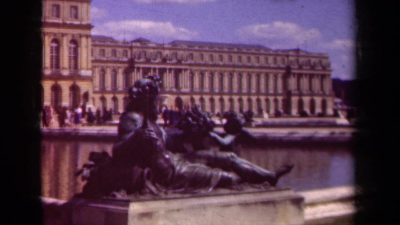 Reclining bronze statue beside a fountain with a palace facade and visitors in the background.
