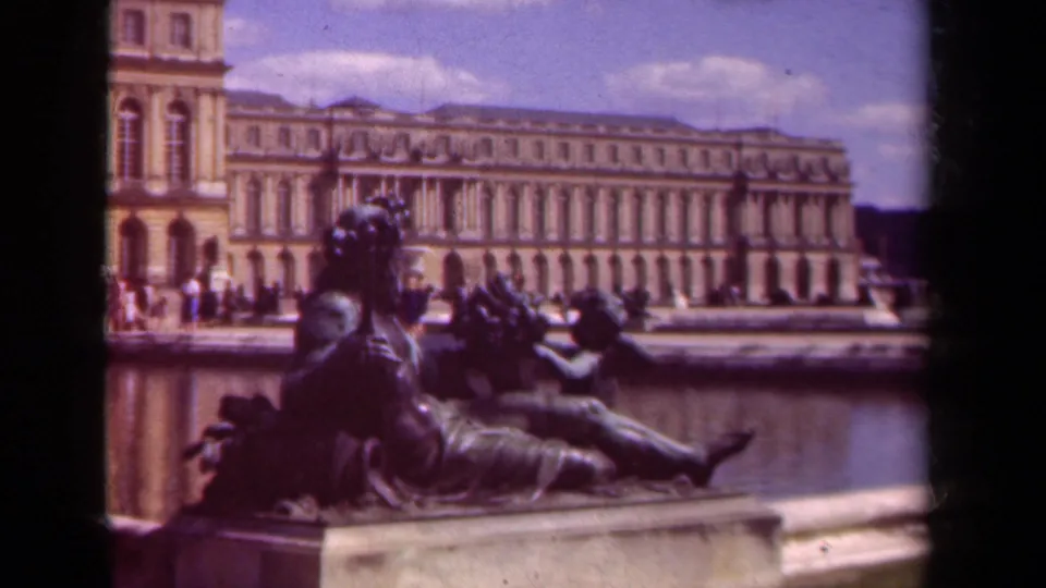 Visitor-shot 8mm frame of a reclining statue by a fountain at Versailles, with the long palace elevation and small figures in the distance.