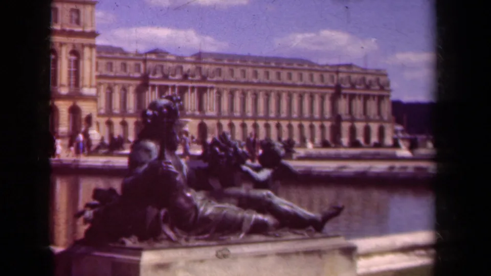 Soft 1939 8mm frame showing the reclining statue and fountain edge before the Versailles palace facade, with visitors moving along the terrace.
