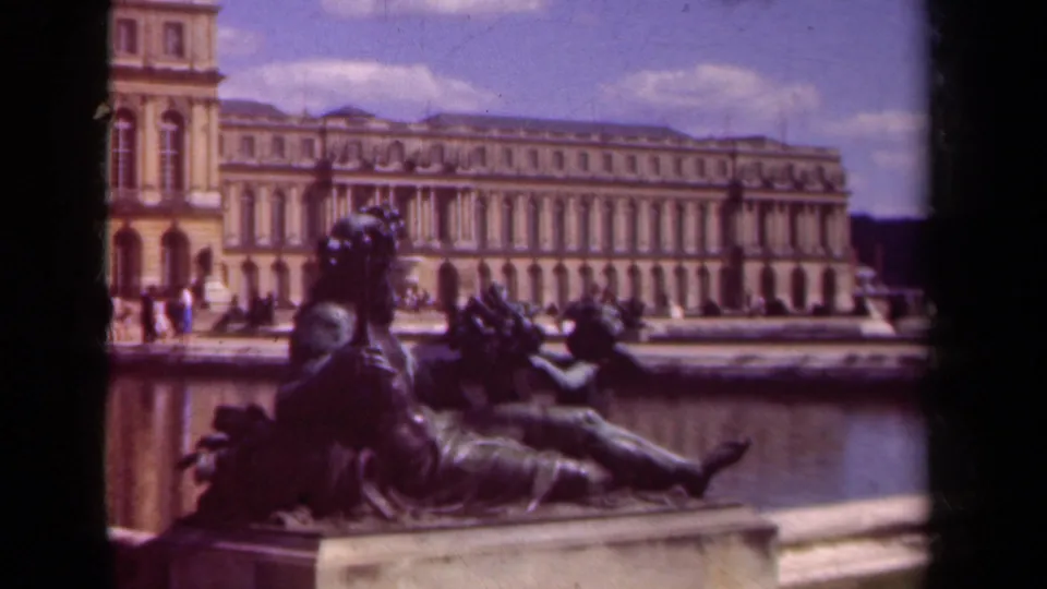 Final 8mm frame of the Versailles fountain area, with a reclining sculpture in front of the palace and small visitor figures along the terrace.