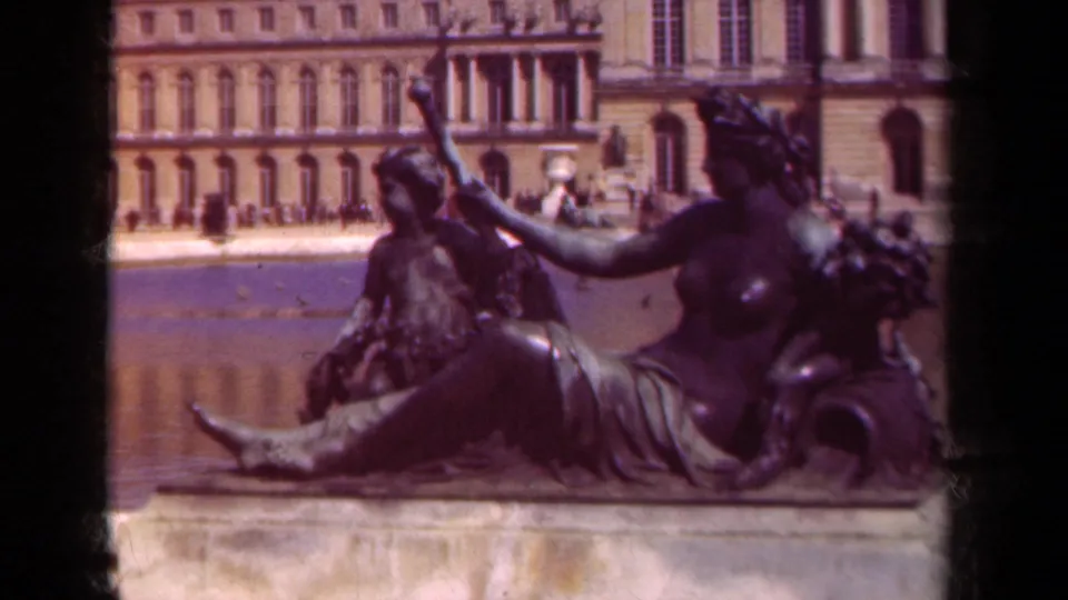 1939 8mm frame showing a foreground classical statue beside the reflecting pool, backed by a formal French palace facade.