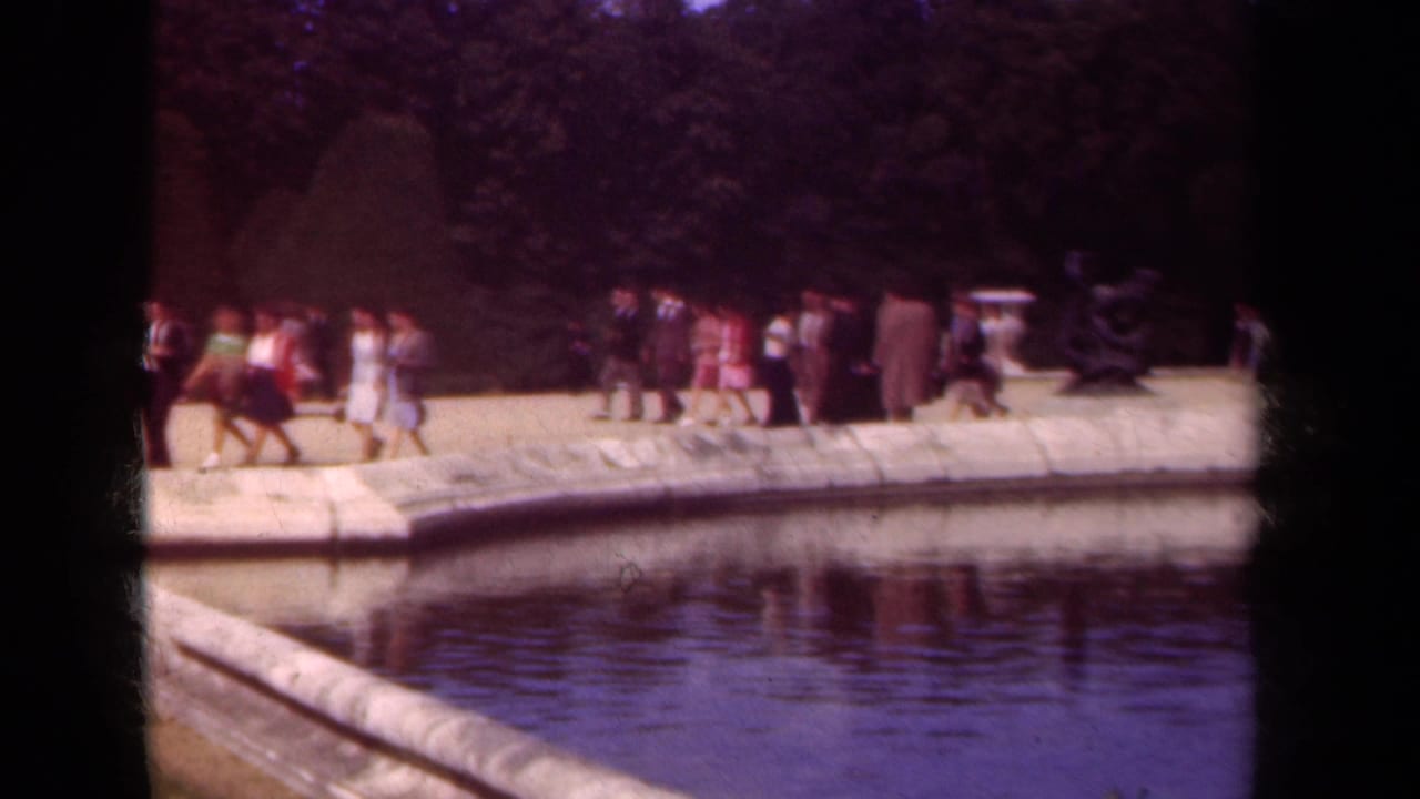 Visitors walk along a curved stone pond edge in a formal park with trees and statuary.