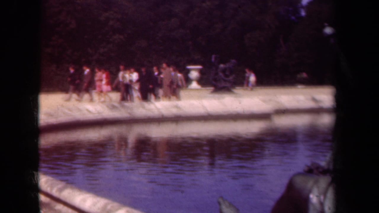 1939 Paris park view with small groups of visitors walking along the pond, framed by a pale stone edge, urn, and shaded trees.
