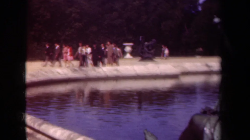 1939 Paris park view with small groups of visitors walking along the pond, framed by a pale stone edge, urn, and shaded trees.