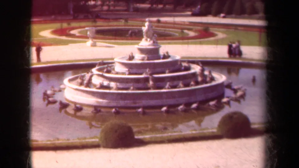 1939 home-movie view of a formal Paris garden fountain with ducks in the basin, circular beds, clipped shrubs, and distant visitors.