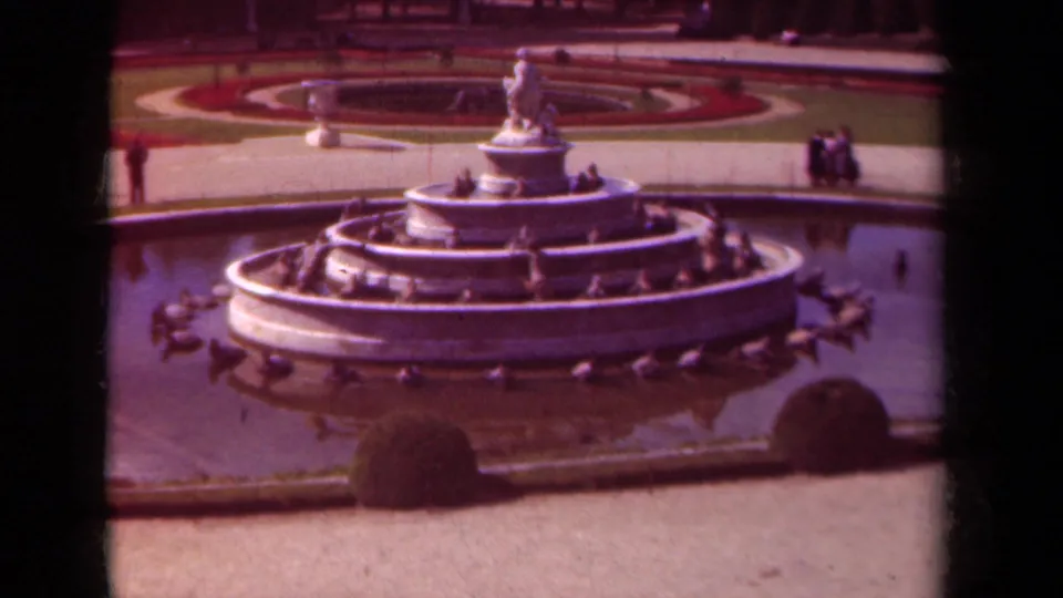 Wide Paris garden view on 8mm film showing a tiered fountain, ducks in the water, circular planting beds, and visitors along the paths.