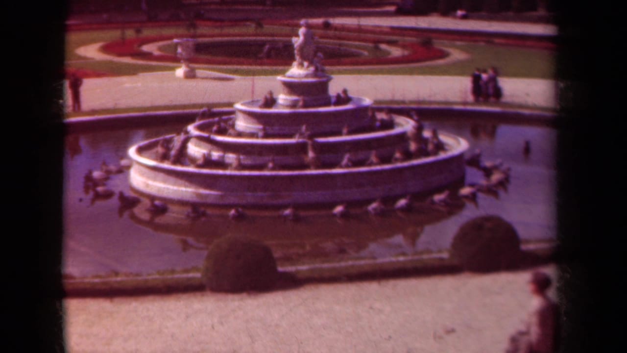 Multi-tiered fountain in a garden with shrubs, paths, and a pedestrian at the lower right.