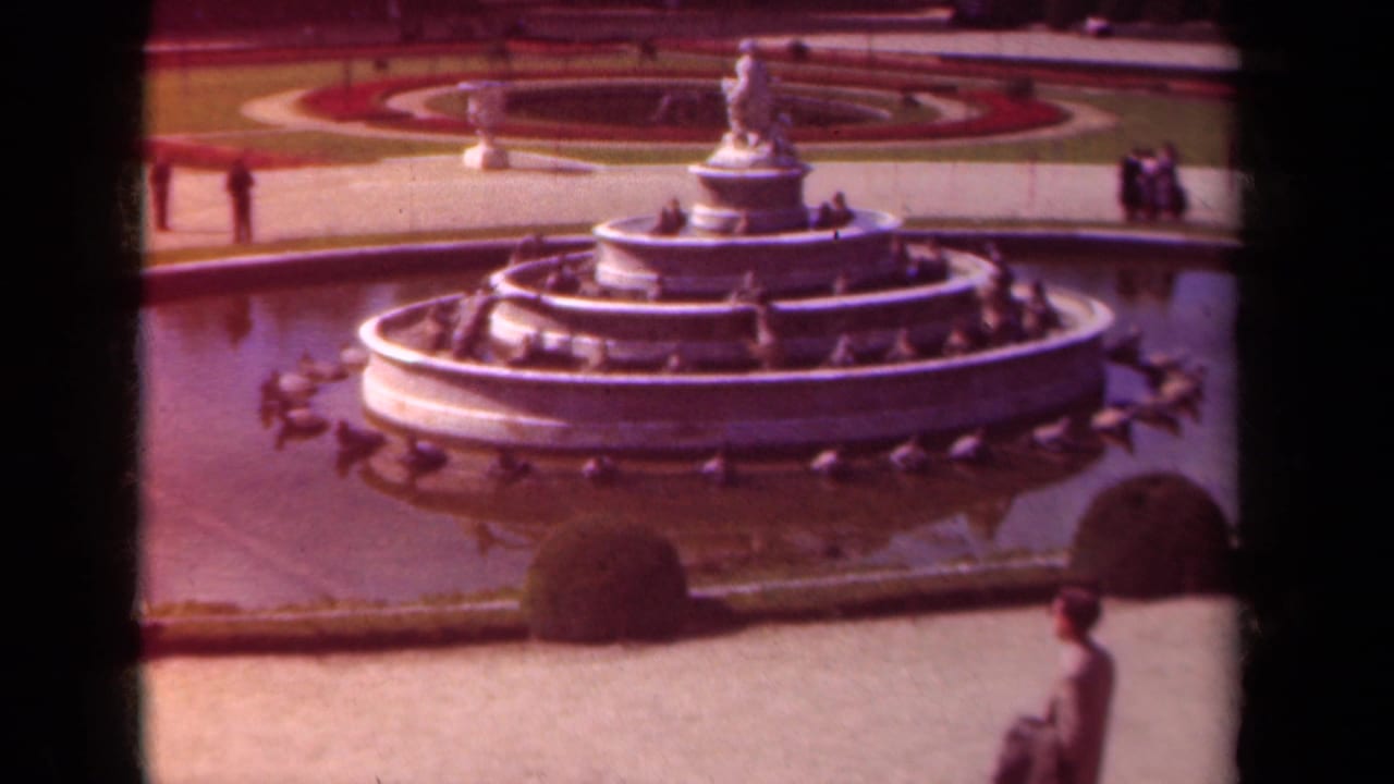 Inactive fountain with circular basin, garden paths, shrubs, and a passerby near the lower right edge.