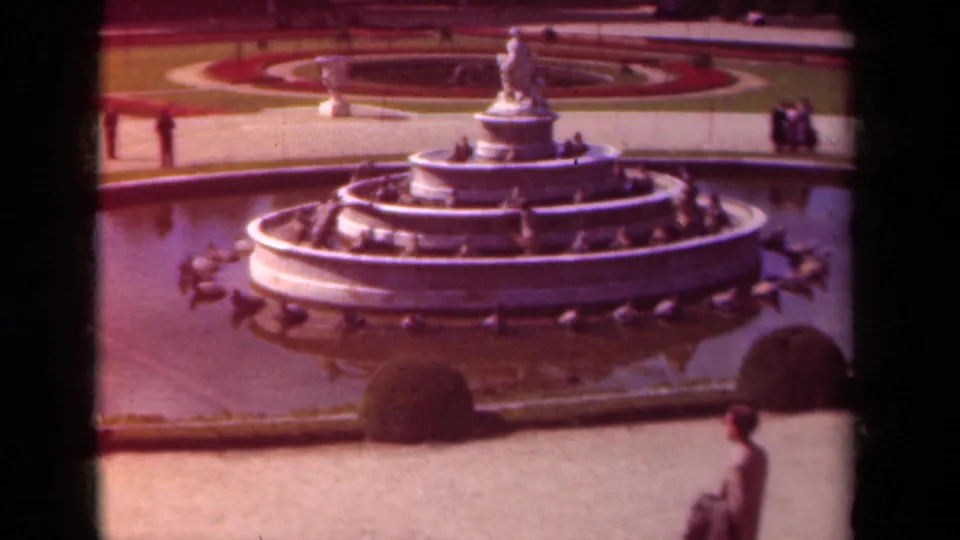 1939 Paris frame with the fountain centered, formal garden paths behind it, clipped shrubs, distant visitors, and a man crossing the foreground.