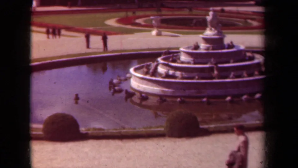 1939 Paris home-movie frame showing the fountain shifted right, basin water in front, formal paths, shrubs, and a person at the lower edge.