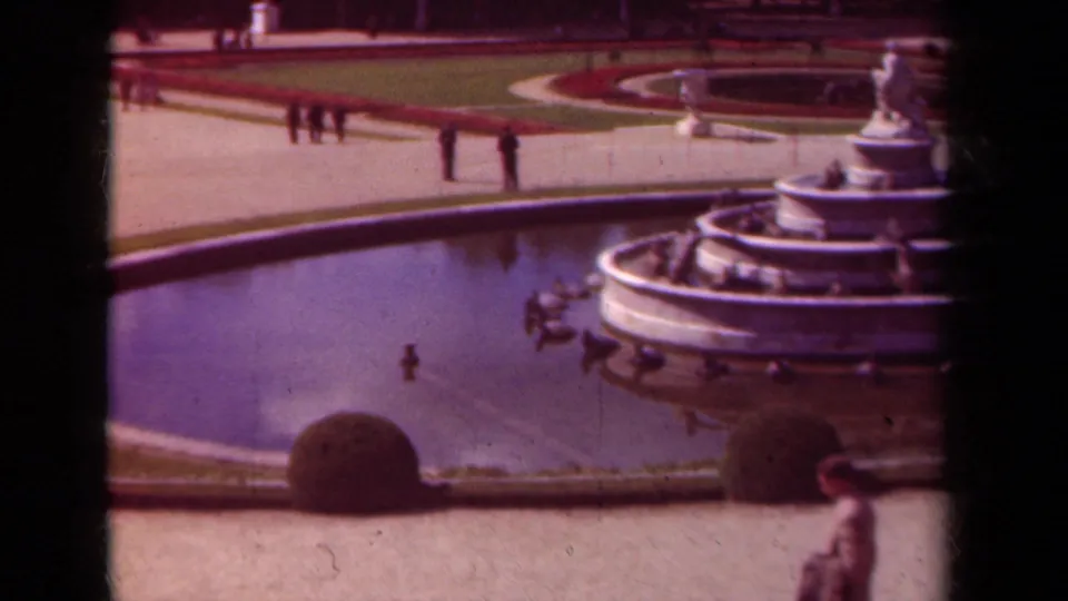 1939 Paris frame showing the fountain partly cropped at right, circular basin, garden paths, distant walkers, shrubs, and a foreground passerby.