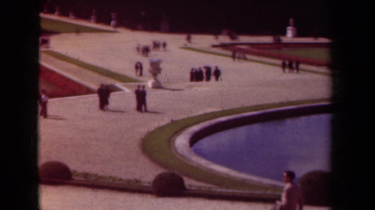 Visitors walk across pale garden paths near a curved pool and red flowerbeds.