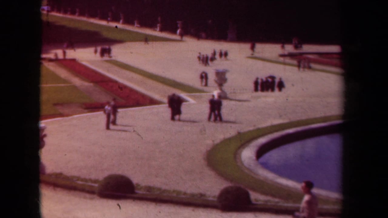 Wide 1939 Paris garden frame showing visitor groups, pale paths, clipped lawns, a white urn, red flowerbeds, and a curved pool.