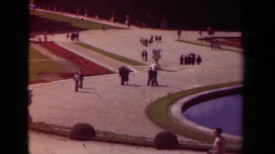 Wide 1939 Paris garden frame showing visitor groups, pale paths, clipped lawns, a white urn, red flowerbeds, and a curved pool.