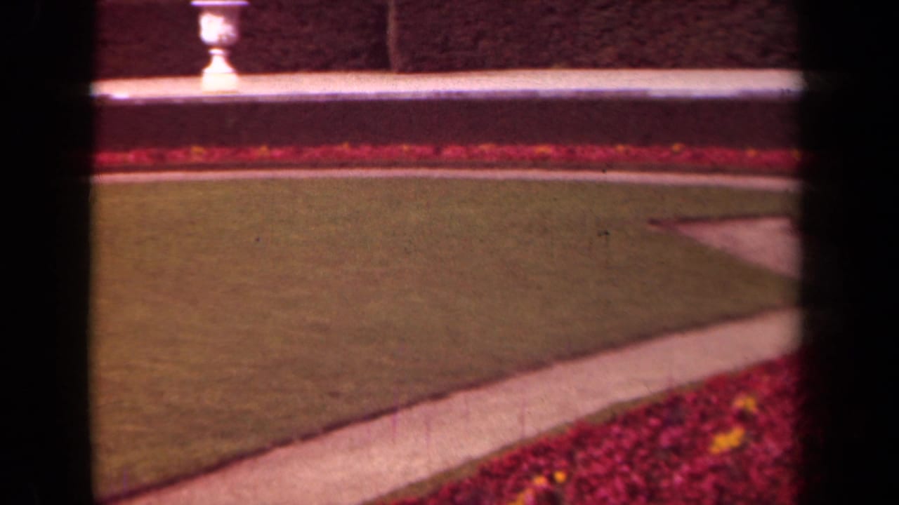 1939 Paris home-movie frame showing a manicured lawn, red flowerbeds, diagonal path, low wall, hedge, and white urn.