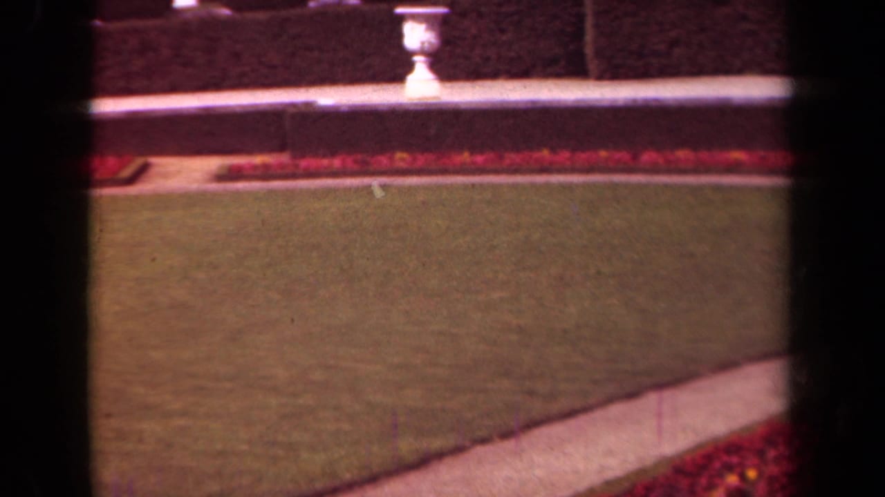 Garden lawn and path bordered by red flowers, with a low wall and white urn beyond.