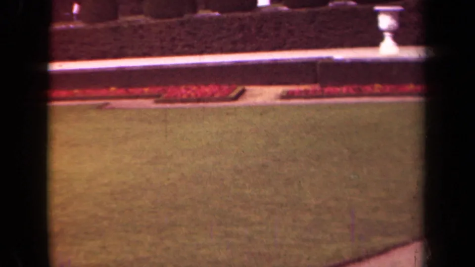 1939 Paris home-movie frame showing lawn, red border plantings, paved path, low wall, hedge, and a white garden urn.