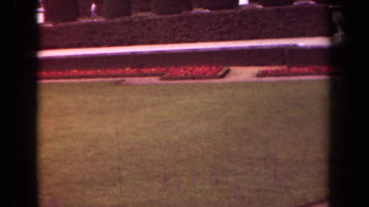 Formal lawn bordered by red flower beds and clipped hedges, seen through dark film gate edges.