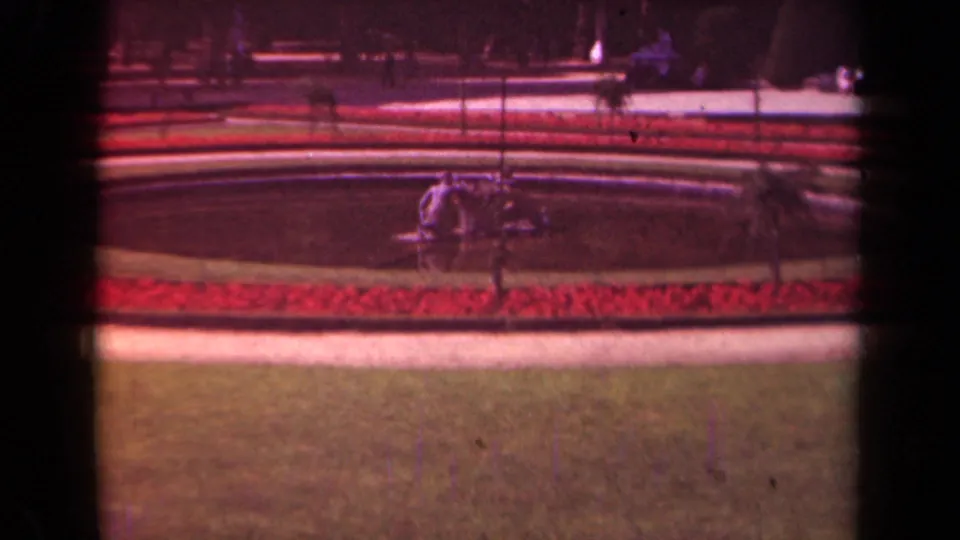 1939 Paris 8mm frame of a zoo attraction area with manicured grass, red flowerbeds, curved paths, railings, and tiny visitors.
