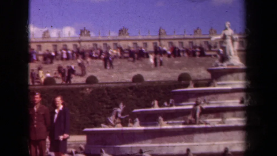 A man in a military-style uniform and a woman in dark clothing stand near the fountain as visitors cross a formal French garden terrace.