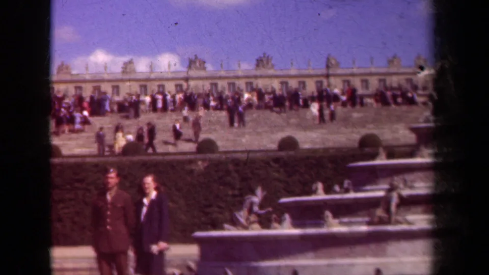 Tourists in period dress move through a broad palace garden terrace, with a stone fountain basin and statues in the foreground.