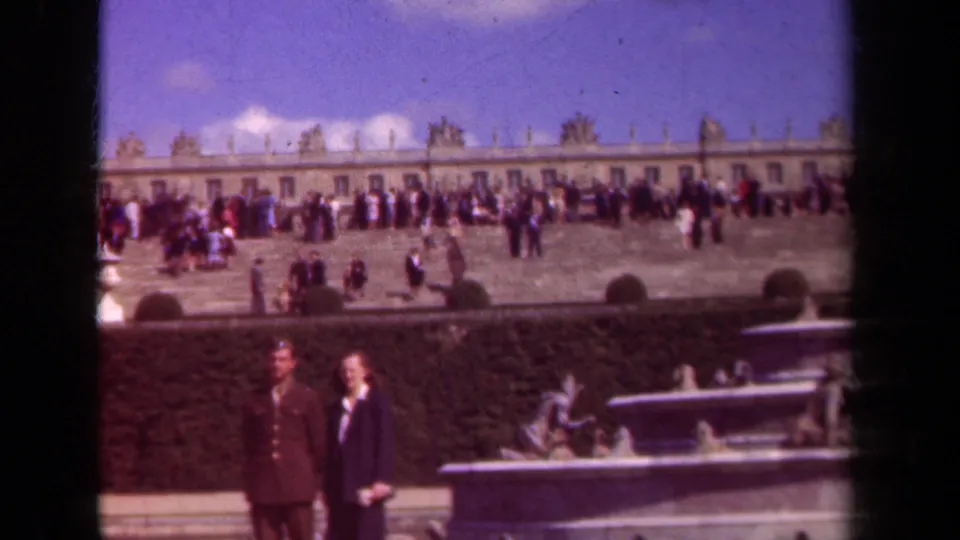 Final frame of the 1939 palace garden view, with tourists on the terrace, fountain sculpture below, and a long formal facade behind them.