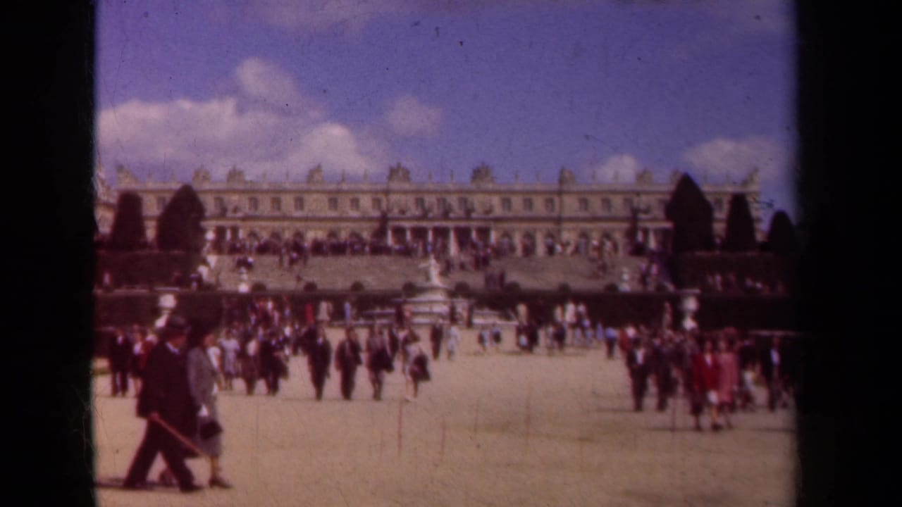Tourists walk across palace grounds with a long facade and formal garden visible behind them.