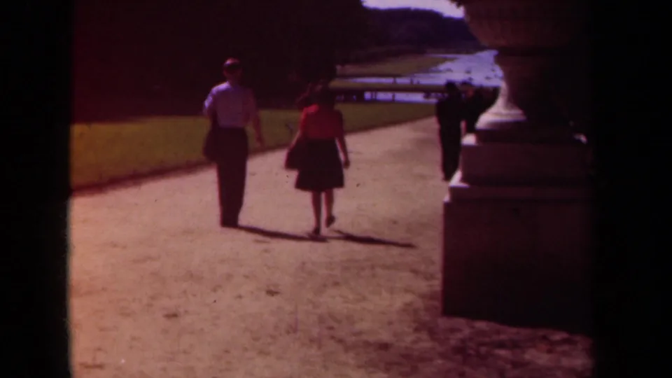 1939 Paris home-movie view of two visitors walking away along a palace garden path near grass and stone ornamentation.