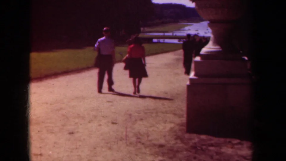1939 Paris frame showing visitors walking through palace gardens, with strong sunlight, shadow, lawn, and stone garden structure.