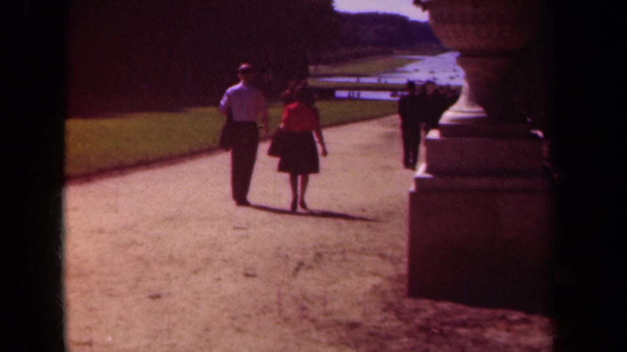 Couple walking hand-in-hand on a sunny gravel path beside a large stone urn.