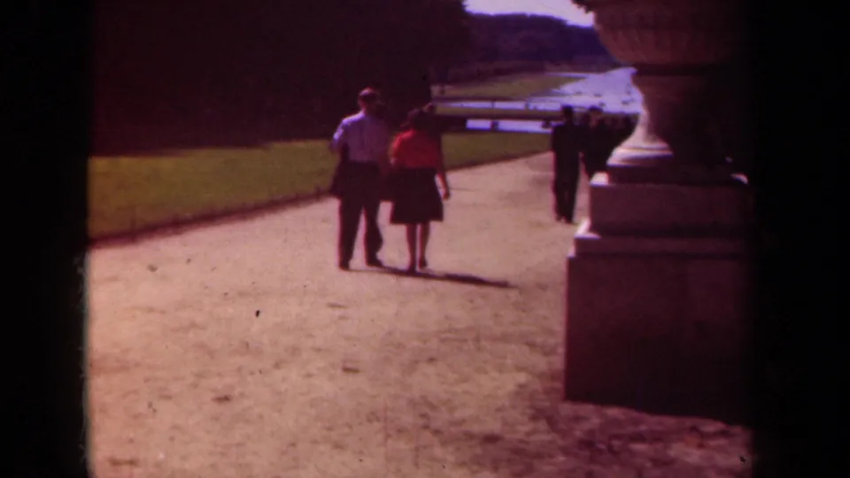 The couple remains centered in the Paris garden scene, walking toward other visitors near a distant water feature.