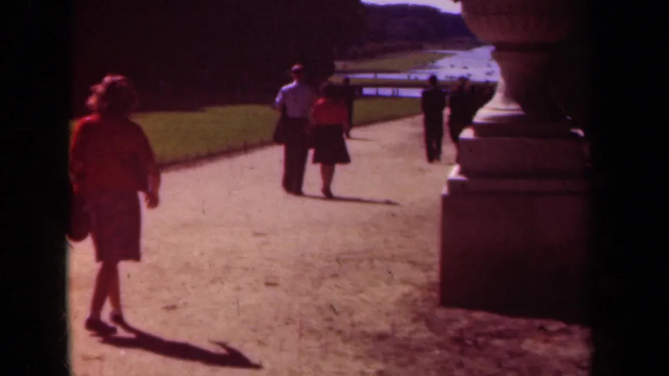 A woman passes close to the camera in the 1939 Paris garden scene as the couple continues down the sunlit path.