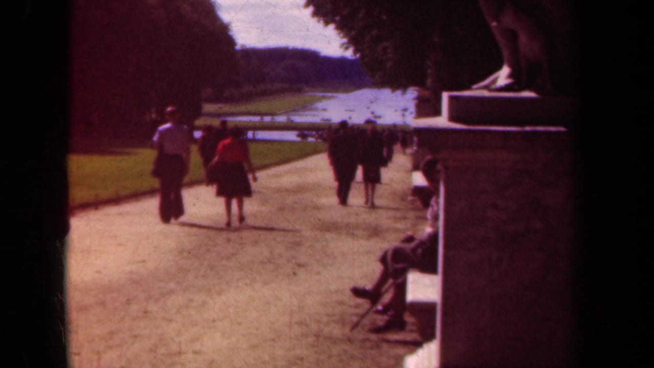 1939 Paris park footage shows adults strolling away along a broad dirt path near lawns, trees, and a long water feature.